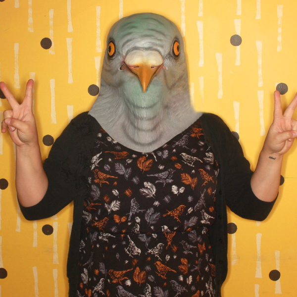 Deanna Seymour in a weird bird mask giving two peace signs on a yellow background with purple dots