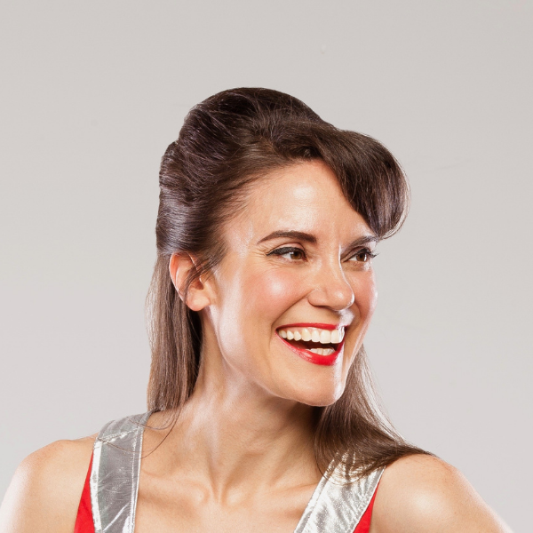 Cassie Stephens with red lipstick and a big smile looking to her left in a silver and red top on a grey background