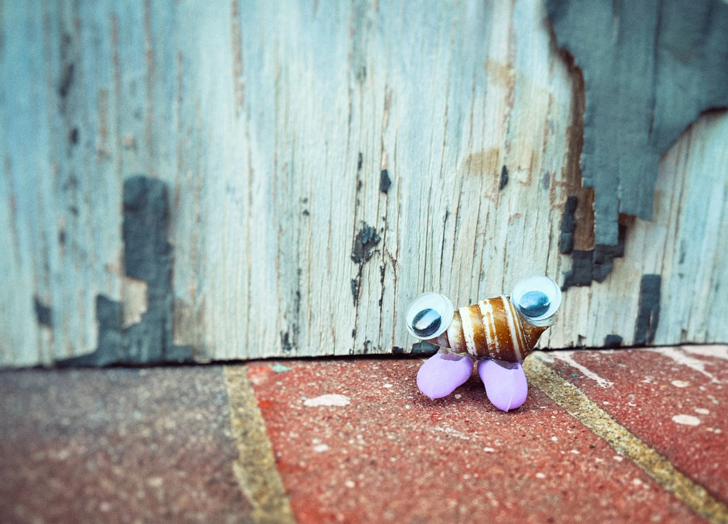A small seashell with google eyes and purple plastic shoes glued onto it standing on some bricks next to some grey rusted and peeling paint.