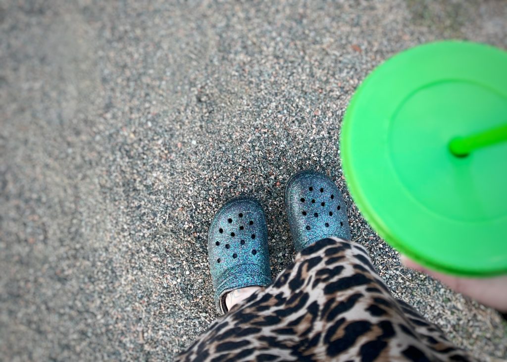 Sparkly crocs, leopard shirt, and green iced coffee cup with straw. Standing on a gravel road with tiny pebbles. 