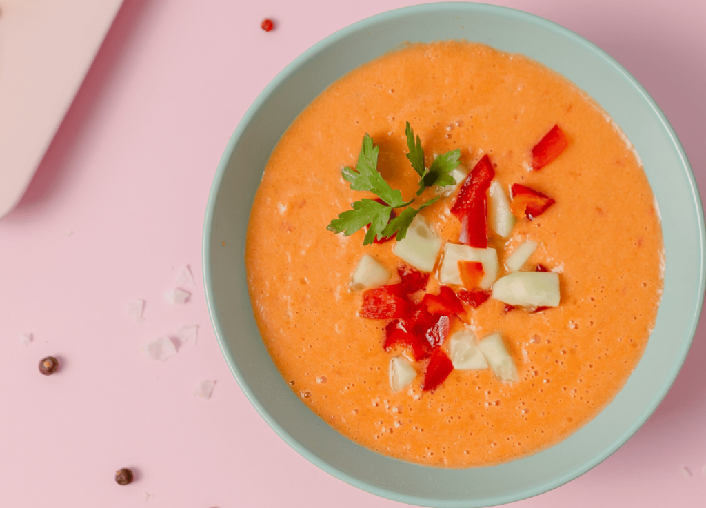 Gazpacho soup topped with parsley onion and peppers in a blue bowl against a pink background