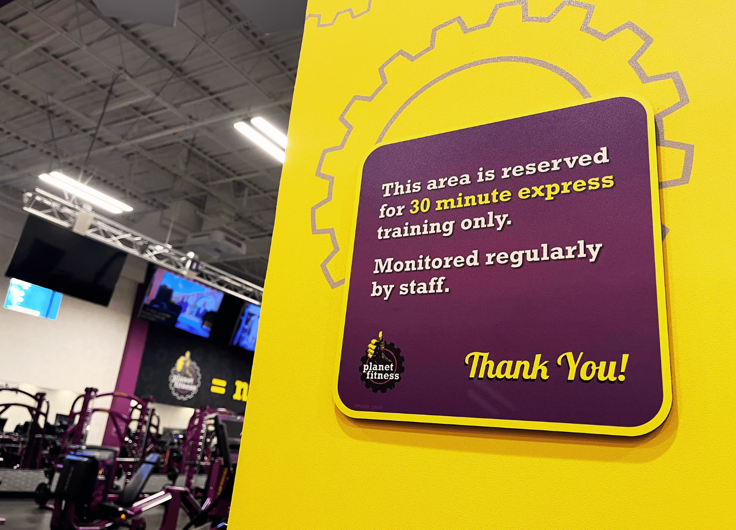 The image shows a sign posted in what appears to be a gym or fitness center. The sign has a yellow background with a gear-like design and the text "This area is reserved for 30 minute express training only. Monitored regularly by staff. Thank You!" There appears to be fitness equipment visible in the background behind the sign.